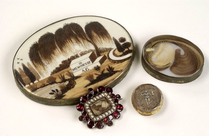 Pieces of Victorian morning jewellery: two brooches and a trinket box decorated with human hair and red gemstones. One of the brooches has a tiny figure of a skeleton.
