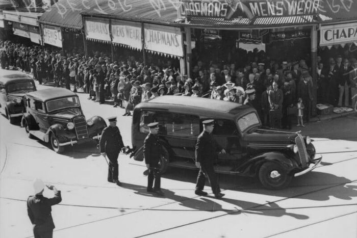 A motor car leads a funeral procession, watched by hundreds