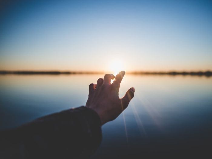 Picture of low sun above a lake with an arm reaching out from bottom left corner and fingers "touching" the sun